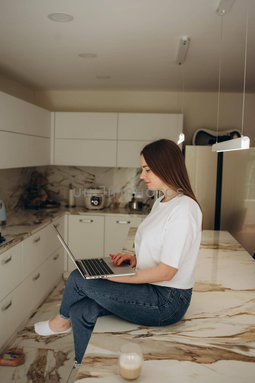 Caucasian female student on video call with friends typing on laptop sipping hot coffee working in kitchen.