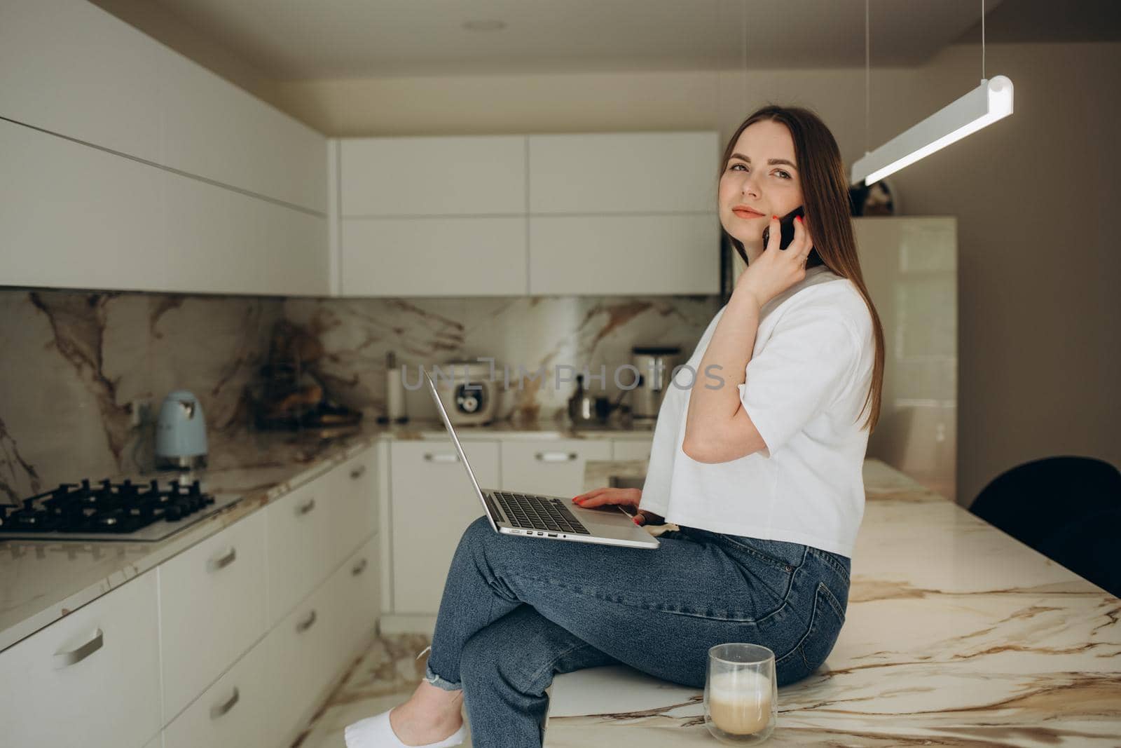 Beautiful girl is talking on the mobile phone, holding a cup, looking away and smiling while sitting on the table in the kitchen. by fentonroma