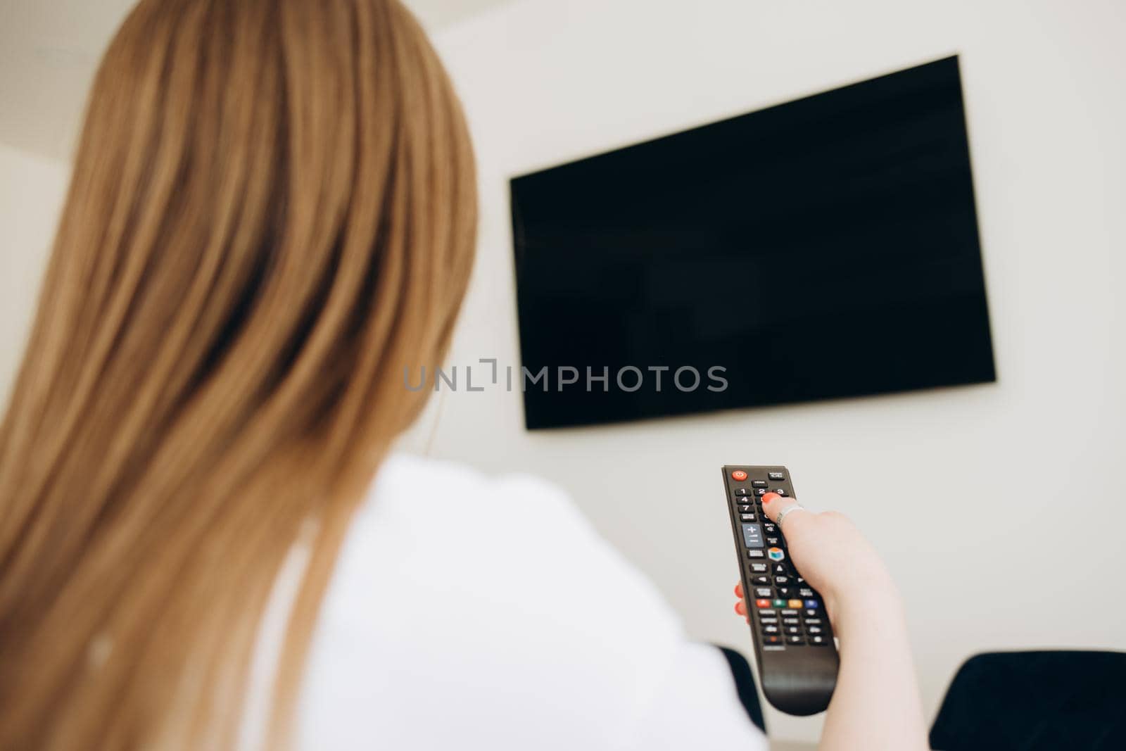 Over the shoulder view of young Asian girl with long black hair sitting on sofa holding tv remote and surfing programs on television. Teenage woman watching TV in the living room by fentonroma