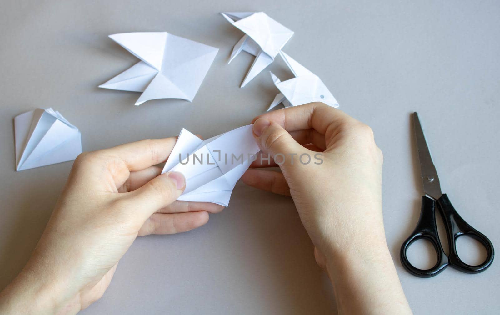 Hands make white paper fish on a gray table.There are scissors next to it.