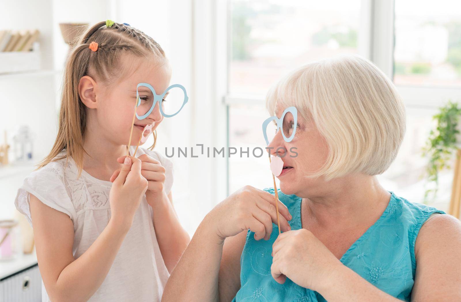 Cute granddaughter and granny holding glasses and lips on sticks in bright room