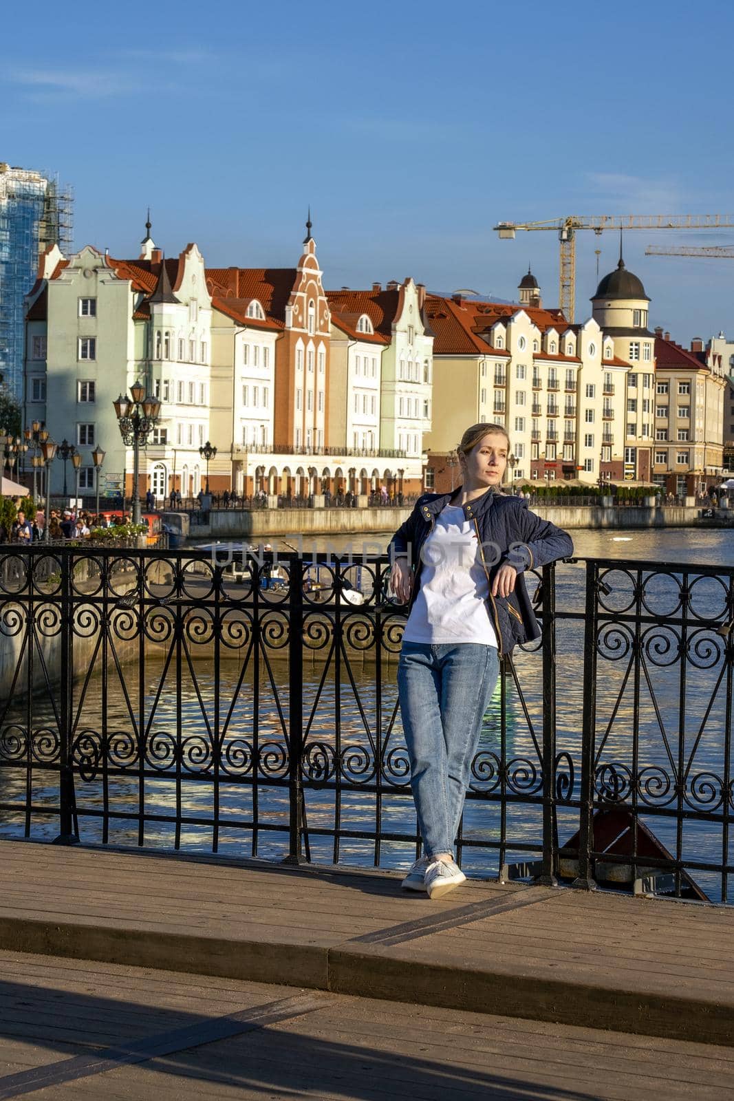 On spring evening, young blonde stands on bridge leaning against railing against backdrop of a beautiful city embankment. Selective focus. Vertical orientation.