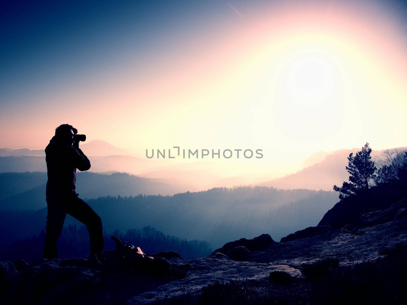 Professional photographer takes photos with mirror camera on peak of rock. Dreamy fogy landscape, spring orange pink misty sunrise in a beautiful valley below