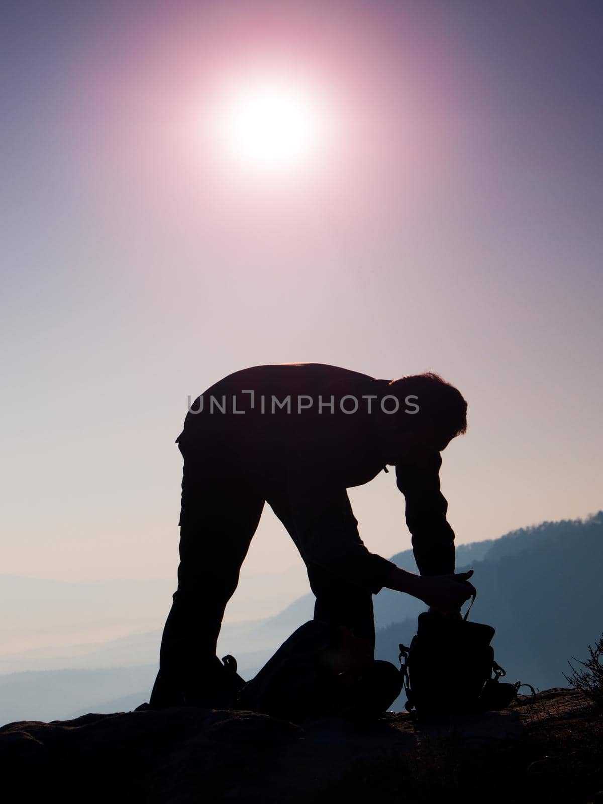 Professional photographer is packing camera into backpack on peak of rock. Dreamy fogy landscape, spring orange pink misty sunrise in a beautiful valley by rdonar2