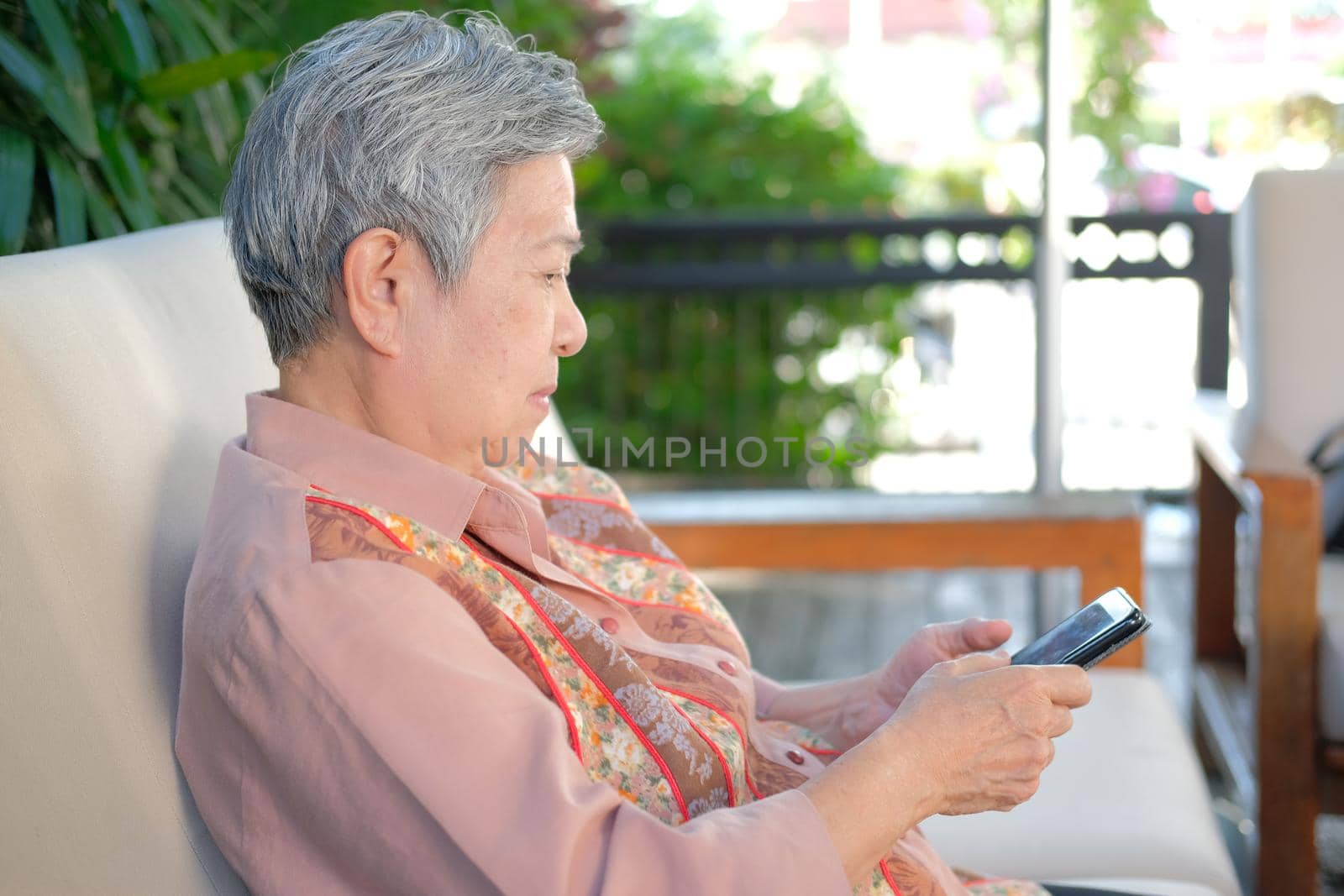 asian elder woman holding mobile phone on terrace. elderly female using smartphone on patio. senior texting message with cellphone