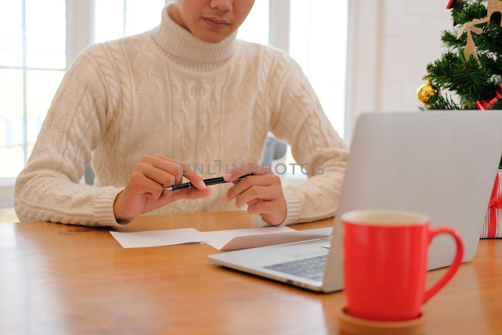 man wearing cream sweater thinking writing christmas letter greeting card holiday wishes with xmas decoration.