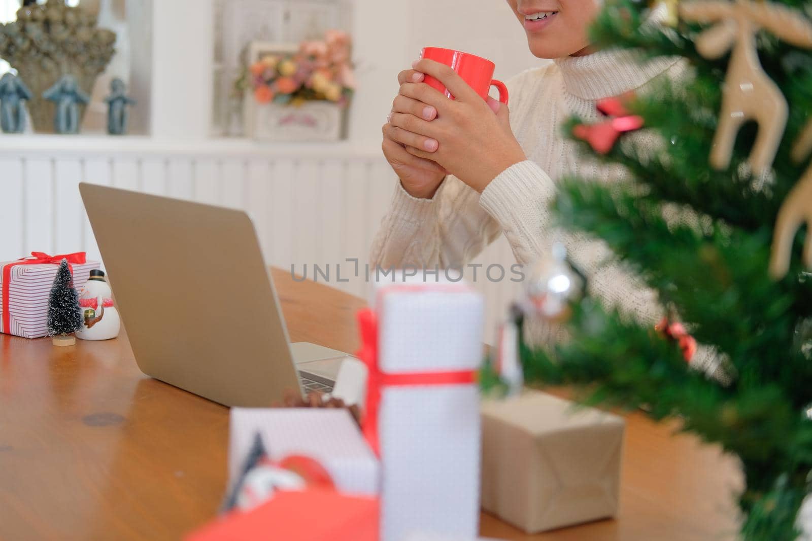 man wearing cream sweater holding coffee cup & using computer at home during christmas. xmas new year celebration