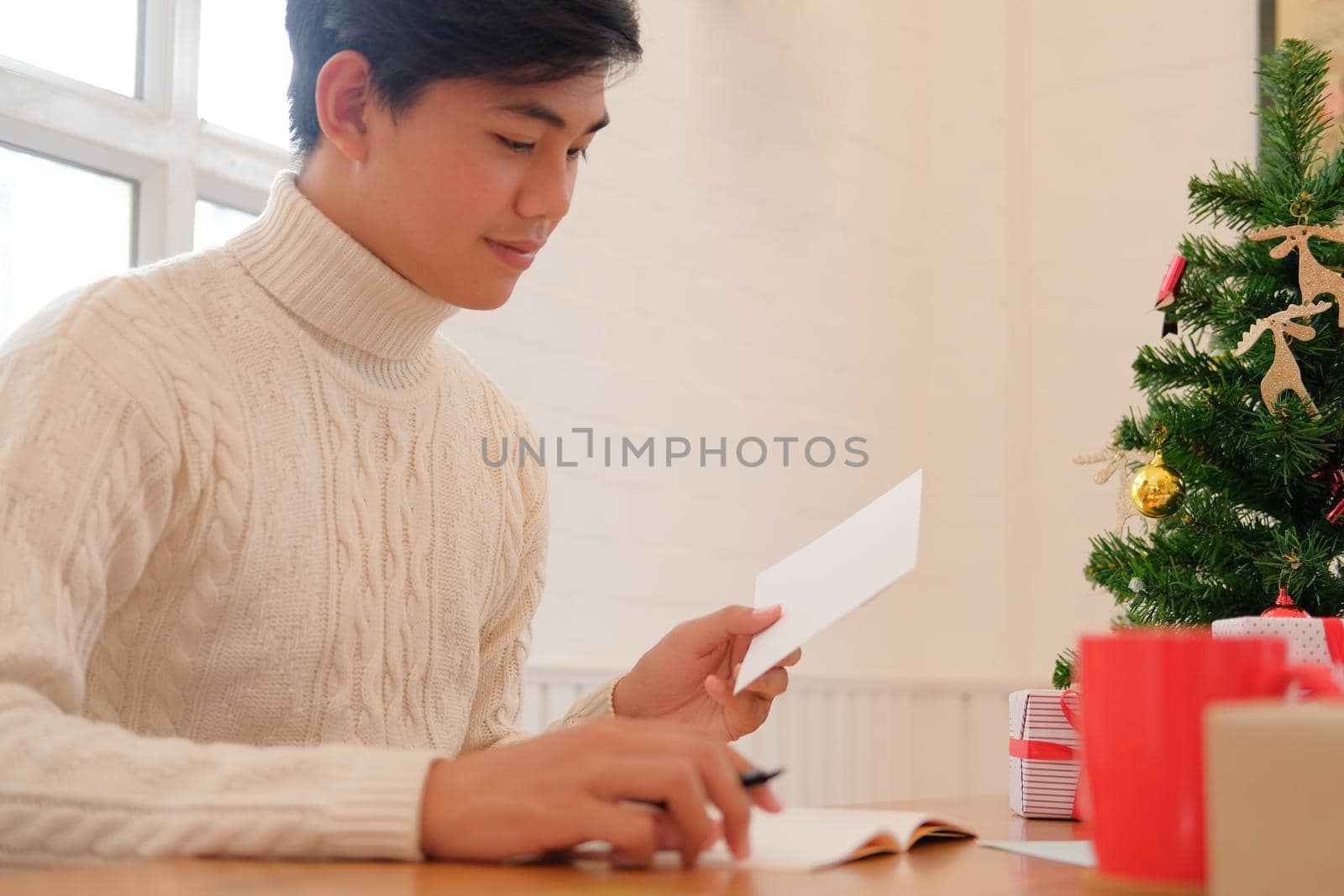 man wearing cream sweater writing christmas letter greeting card holiday wishes with xmas decoration.