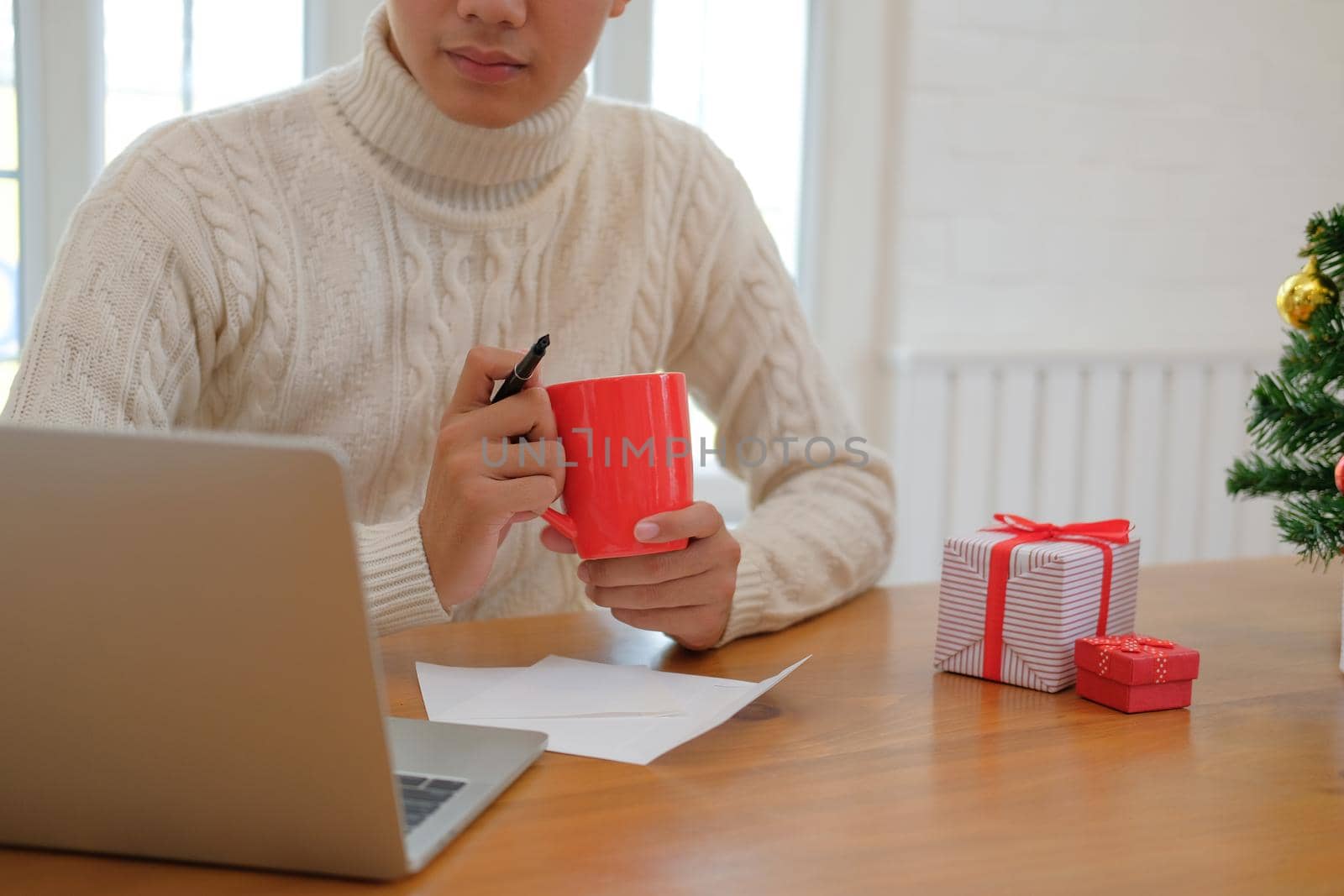 man wearing cream sweater holding coffee cup & using computer at home during christmas. xmas new year celebration