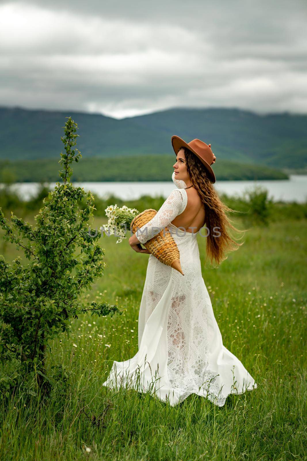 A middle-aged woman in a white dress and brown hat stands with her back on a green field and holds a basket in her hands with a large bouquet of daisies. In the background there are mountains and a lake