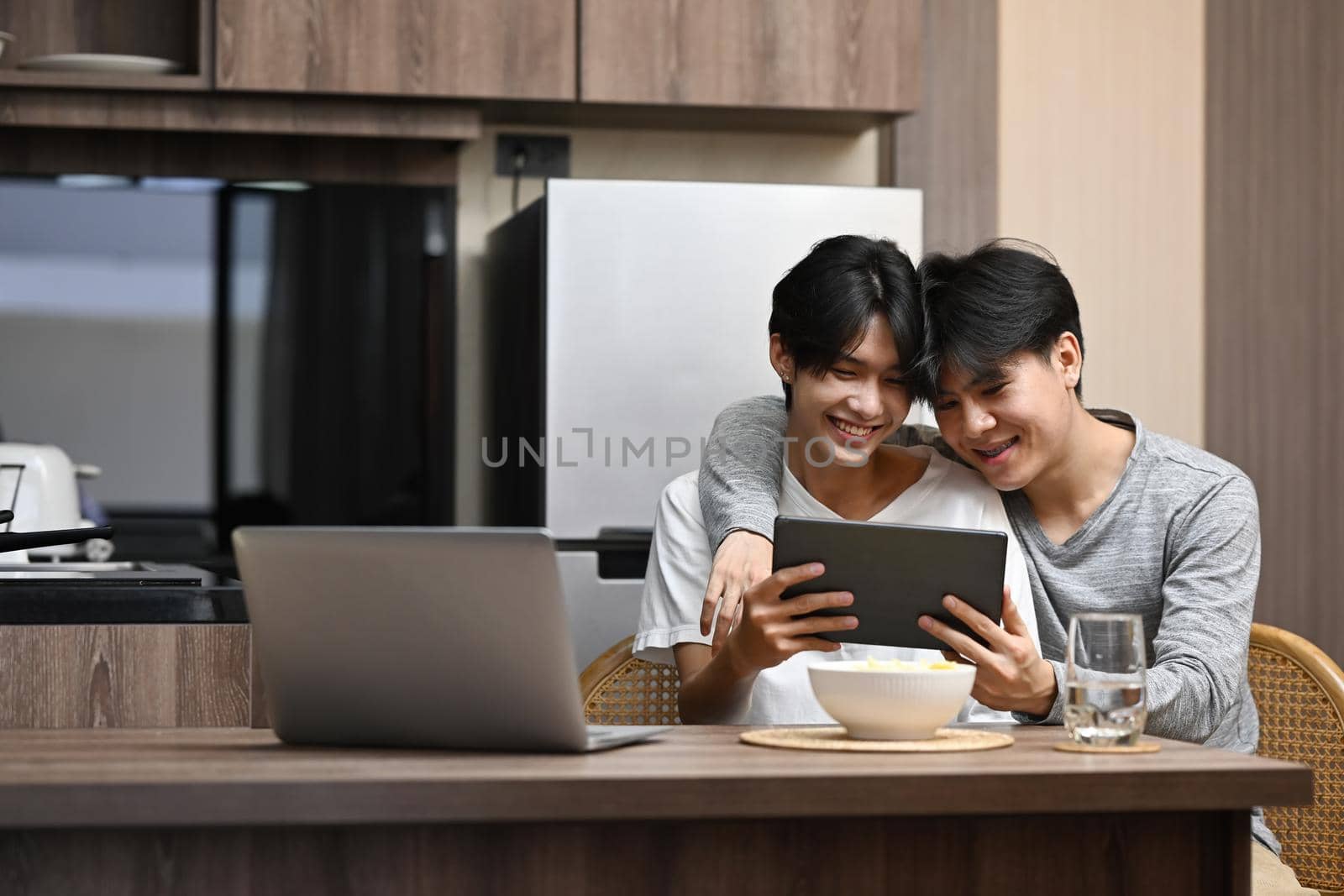 Happy gay couple embracing and surfing internet with digital tablet together at kitchen table. LGBT, pride, relationships and equality concept.