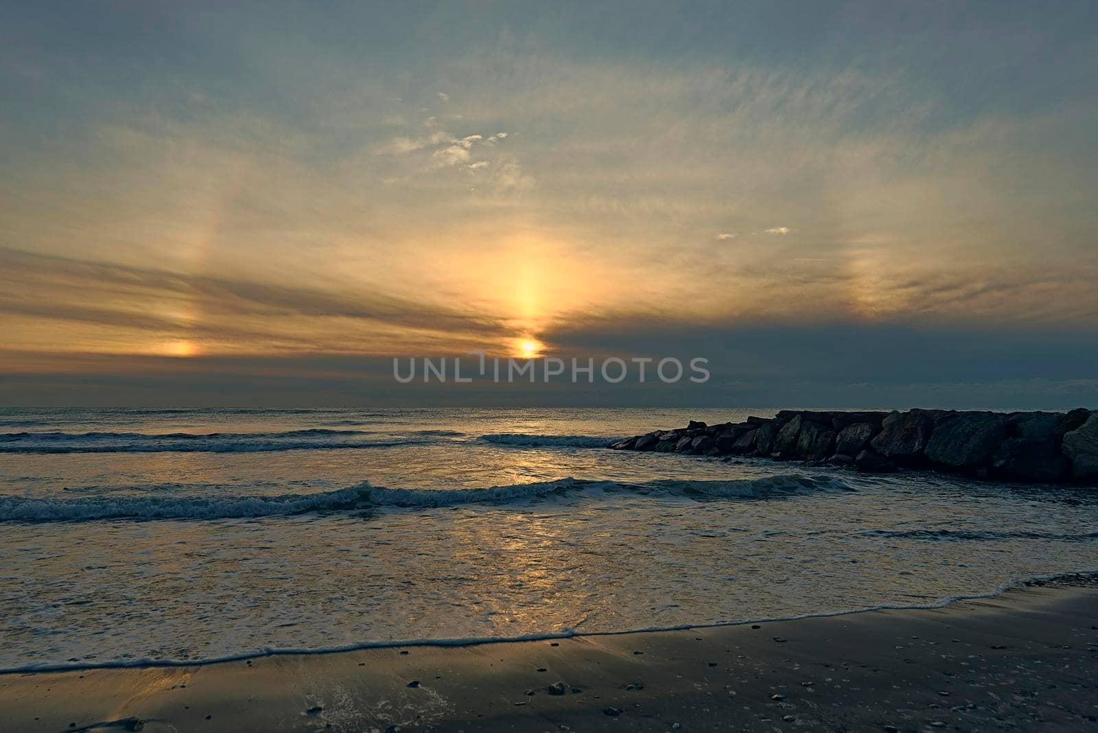 Sunrise from the beach with a breakwater. Rainbow, Circle around the sun, clouds and blue sky, waves, calm sea