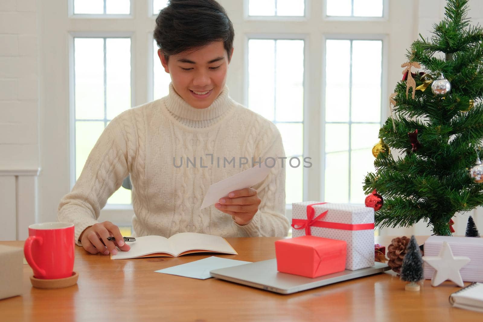 man wearing cream sweater writing christmas letter greeting card holiday wishes with xmas decoration.
