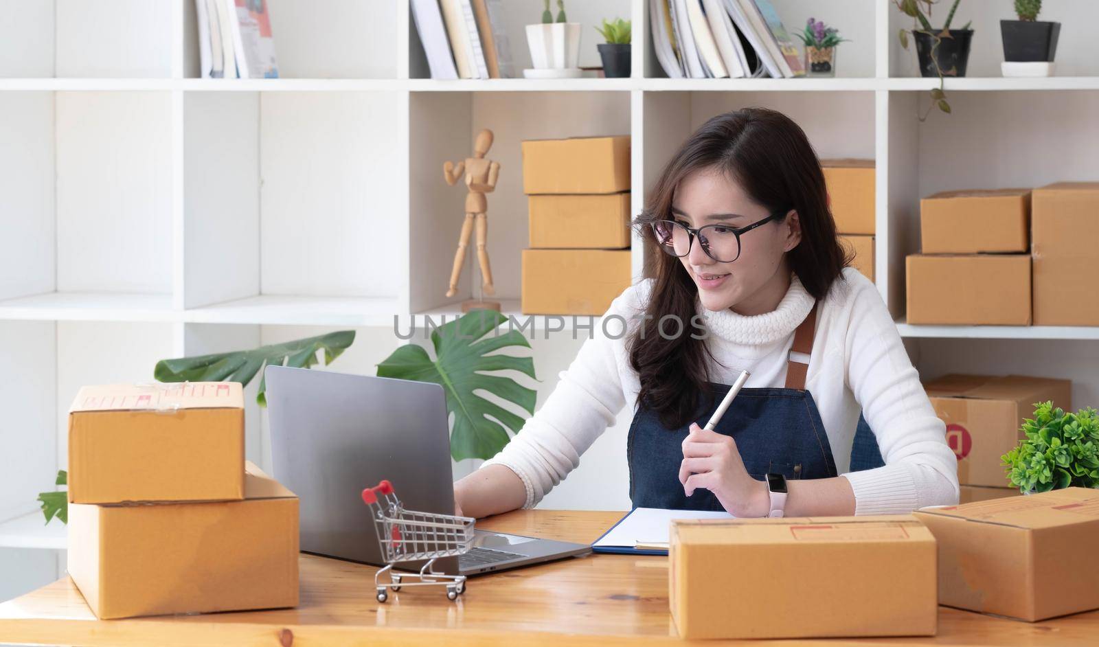 Young beautiful happy asian business woman owner of SME online using laptop receive order from customer with parcel box packaging at her startup home office, online business seller and delivery by wichayada