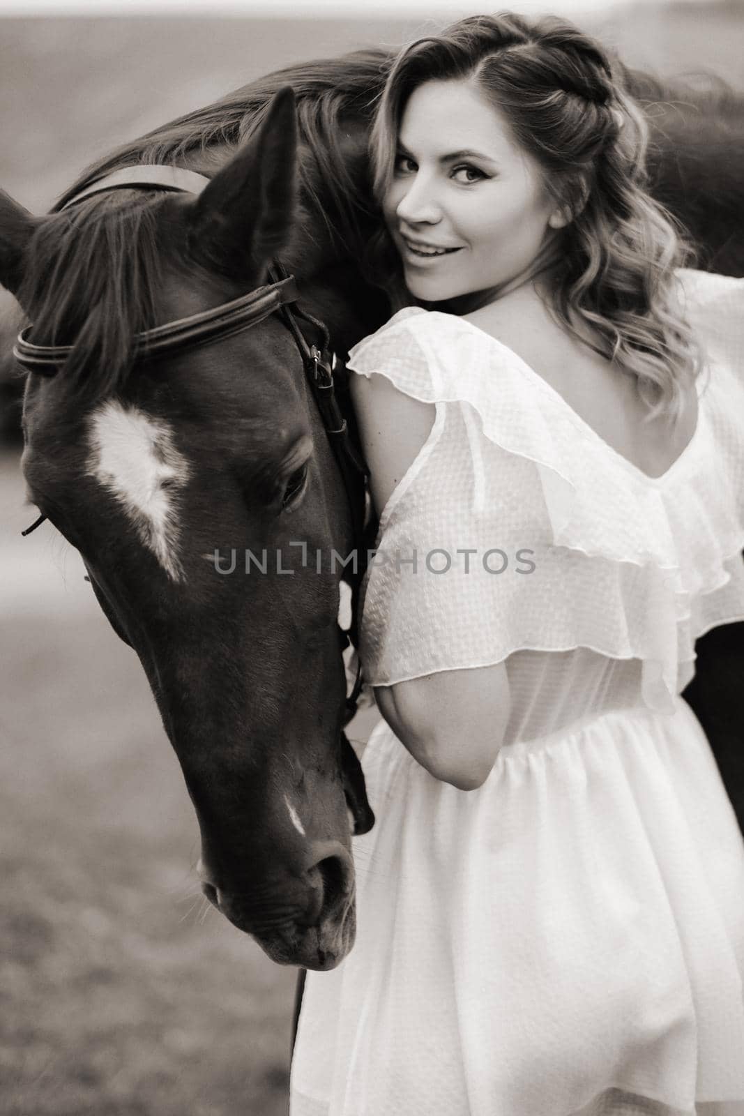 A girl in a white sundress stands next to a brown horse in a field in summer. black and white photo by Lobachad