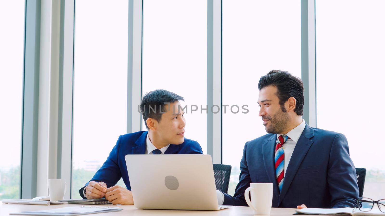 Two business people talk project strategy at office meeting room. Businessman discuss project planning with colleague at modern workplace while having conversation and advice on financial data report.