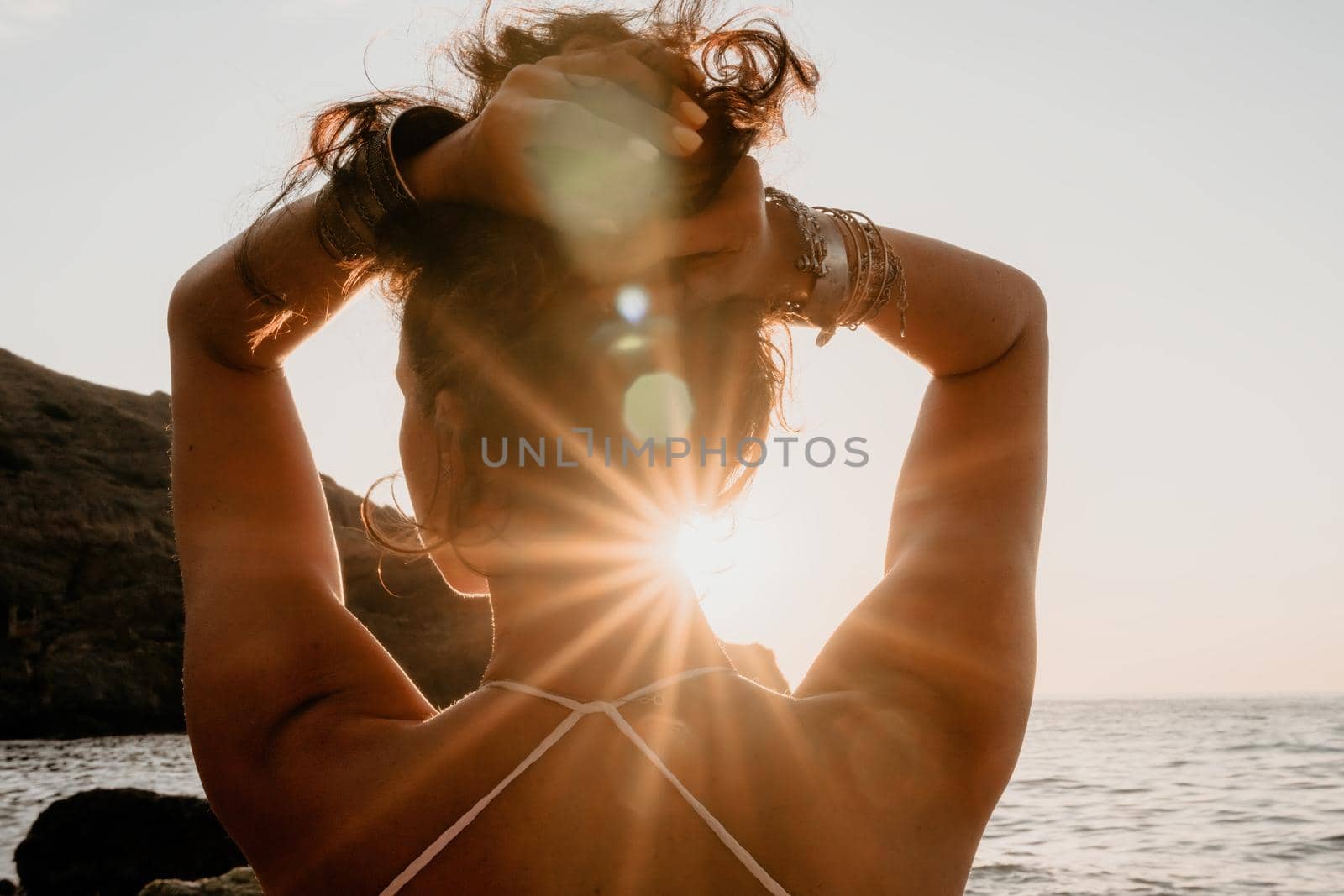 Middle aged well looking woman in white swimsuit and boho style braclets practicing outdoors on yoga mat by the sea on a sunset. Women's yoga fitness routine. Healthy lifestyle, harmony and meditation by panophotograph