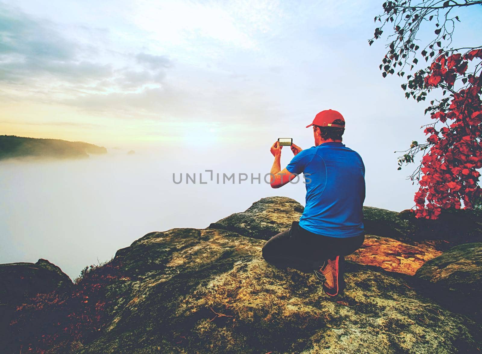 Courageous man sitting on the peak of rock makes the photo on the his phone. Autumn nature 