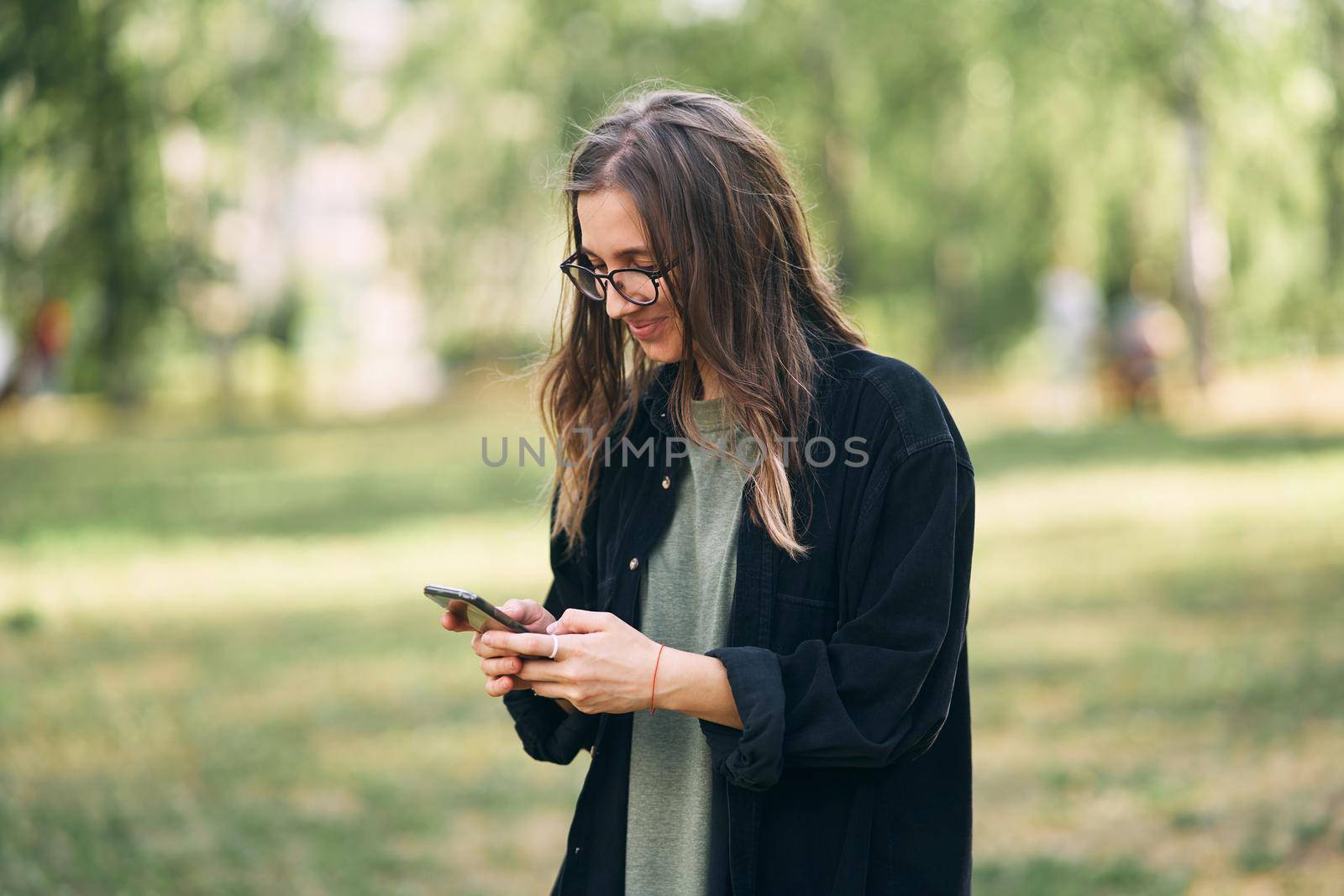 Young woman with glasses reading a message on her phone while standing in the park. High quality photo