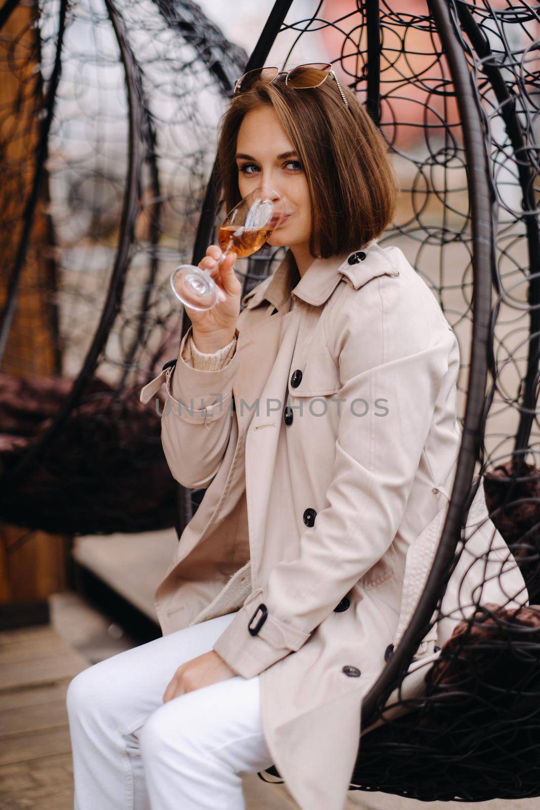 A happy stylish girl In a gray coat is sitting outside in an armchair and drinking a drink by Lobachad