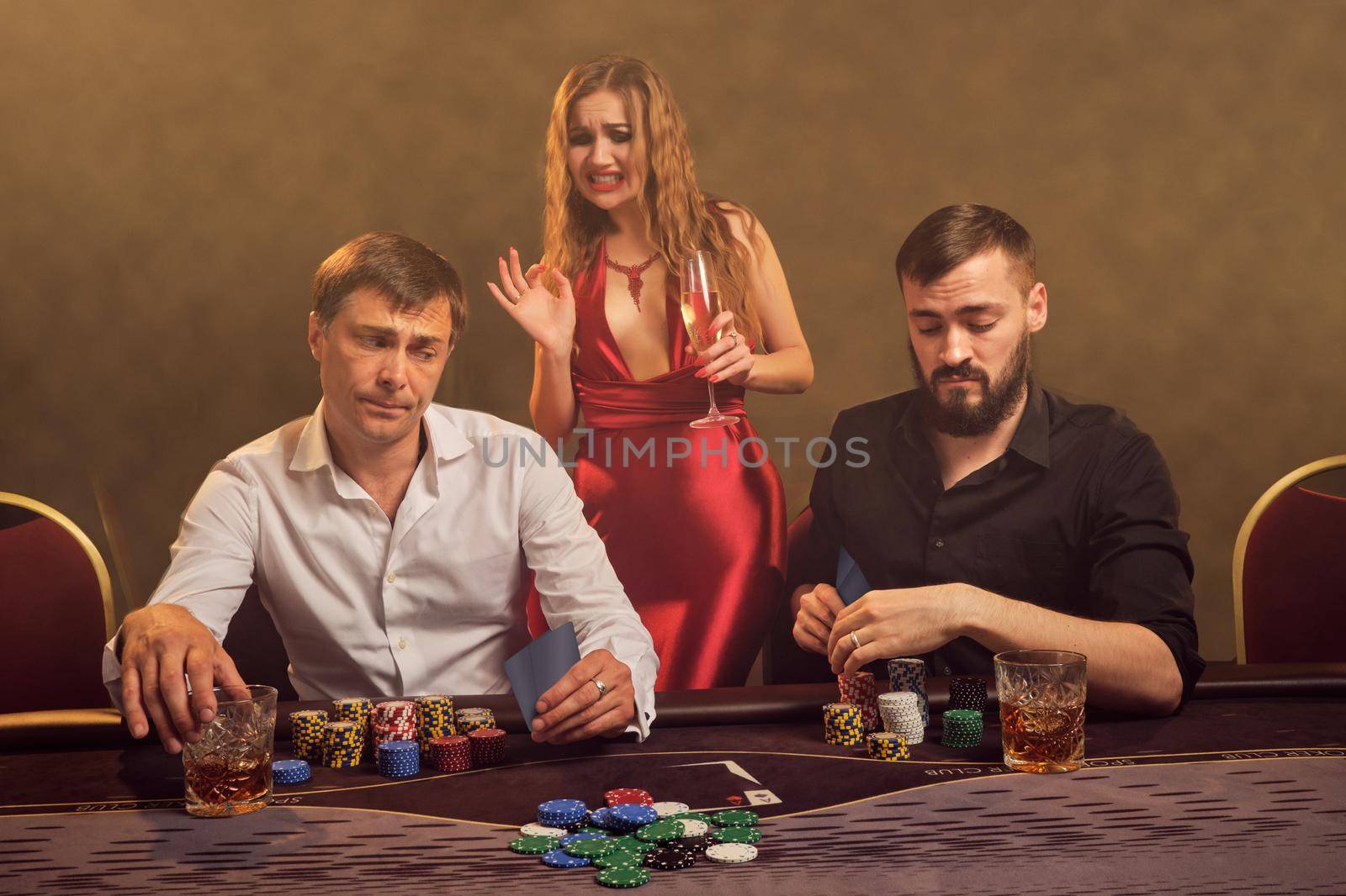 Two old friends and attractive female are playing poker at casino. They are making bets waiting for a big win and looking a little bit disappointed while posing at the table against a yellow backlight on smoke background. Cards, chips, money, gambling, entertainment concept.