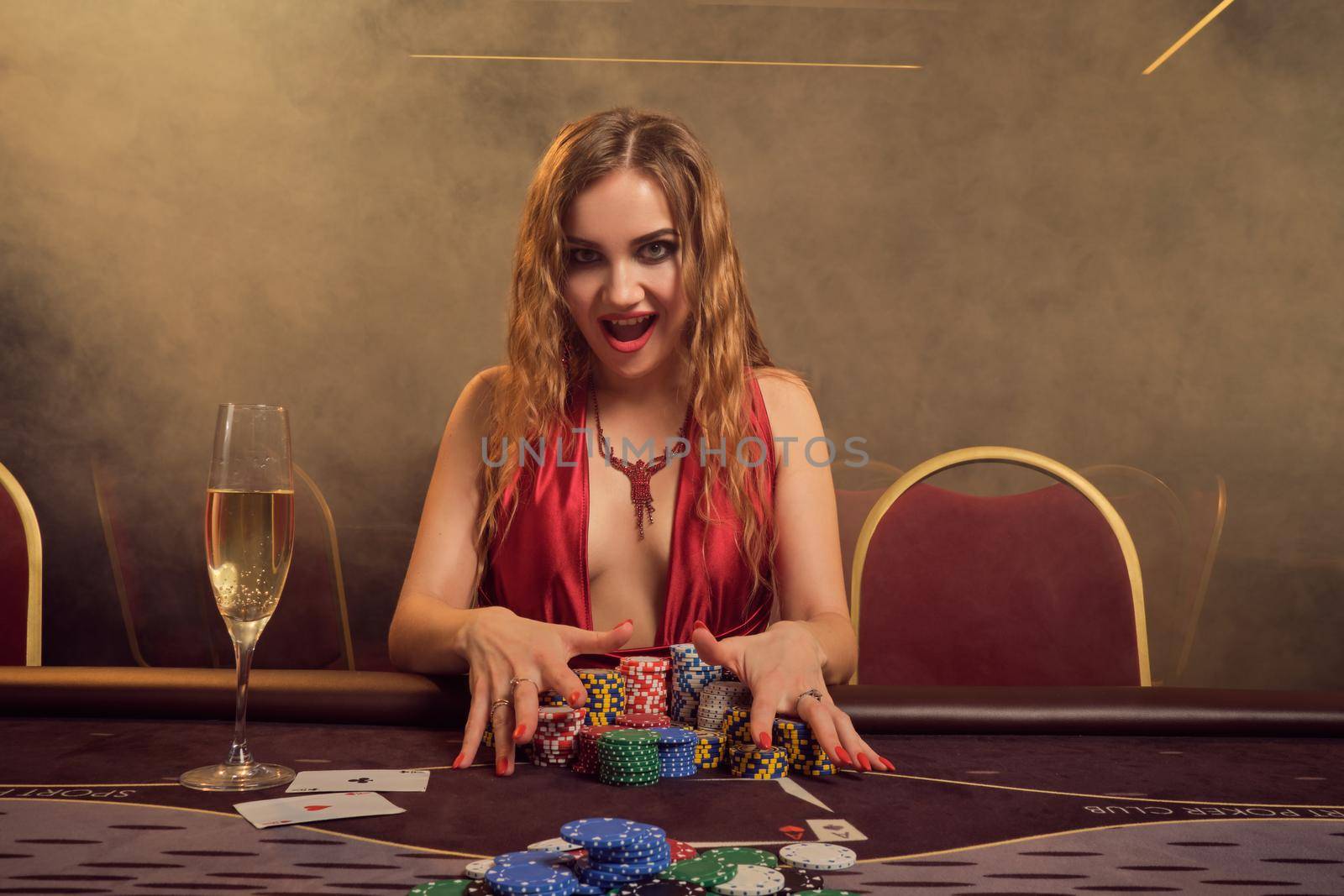 Attractive woman with a long curly hair and perfect make-up, dressed in a sexy red dress. She is sitting at a gambling table, holding some gambling chips and smiling. Poker concept on a dark smoke background in a ray of a yellow spotlight. Casino.