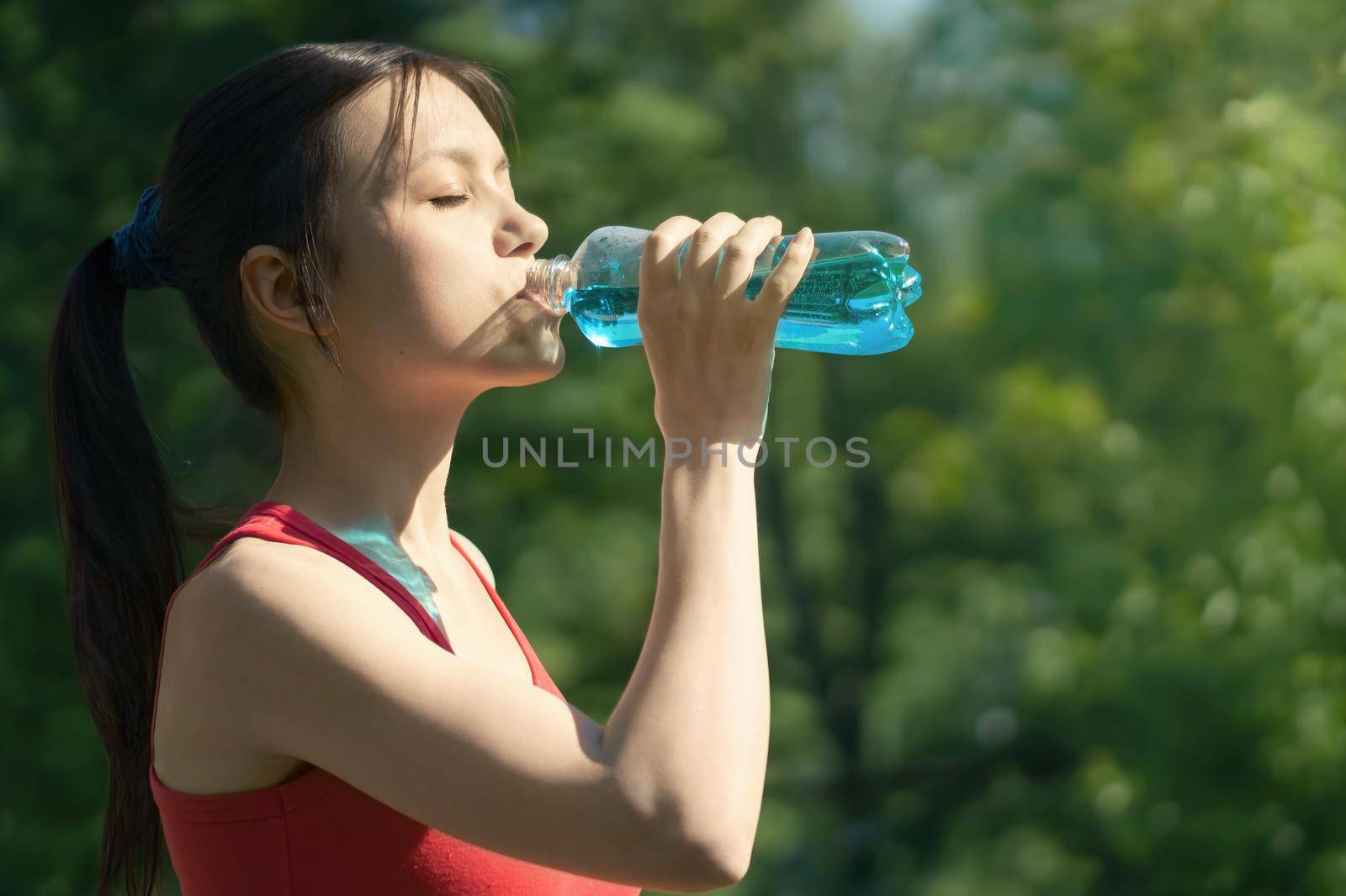 Beautiful young woman jogging in the morning in the park drinking blue isotonic from a bottle.