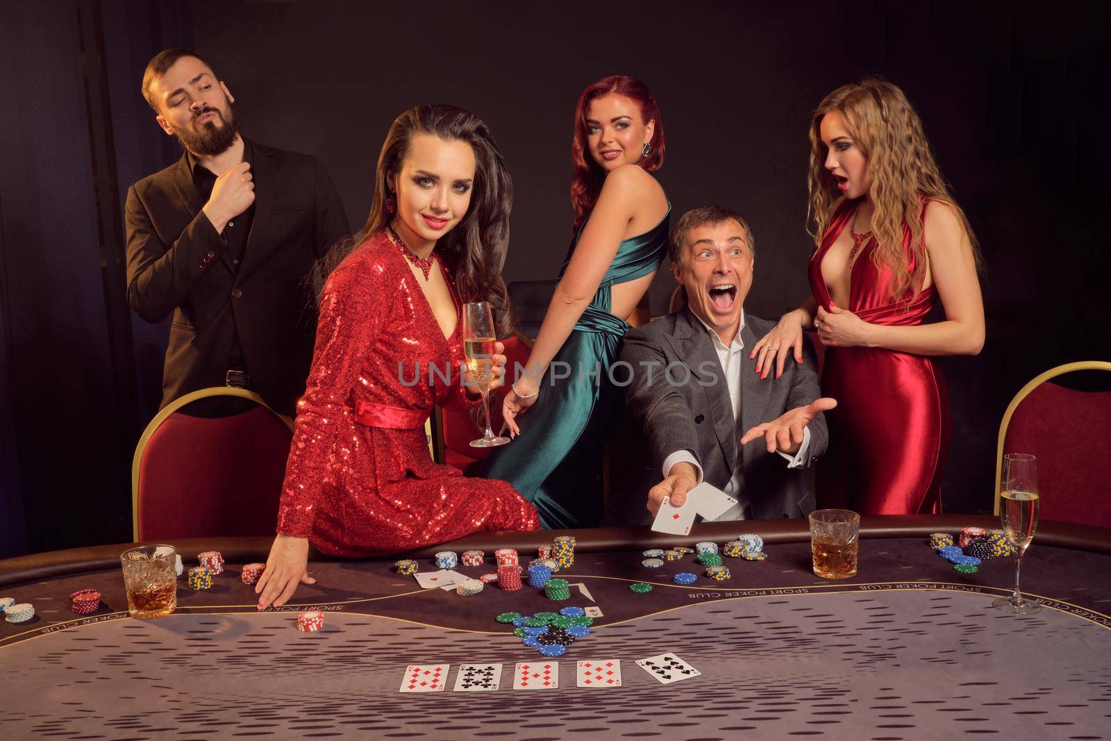 Joyful partners are playing poker at casino. They are celebrating their win, smiling and looking vey excited while posing at the table against a dark background. Cards, chips, money, alcohol, gambling, entertainment concept.