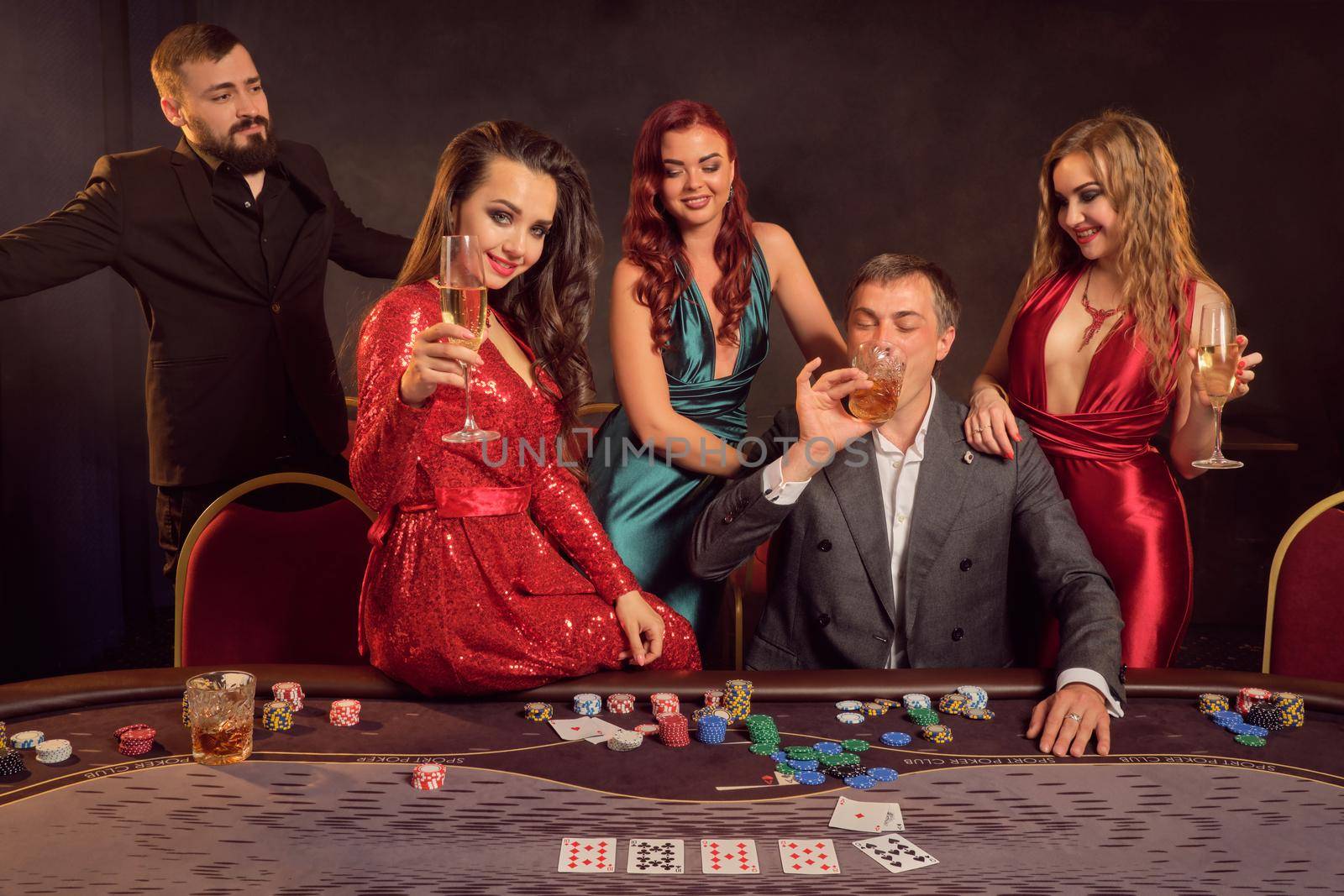 Joyful classmates are playing poker at casino. They are celebrating their win, smiling and looking vey excited while posing at the table against a dark background. Cards, chips, money, alcohol, gambling, entertainment concept.