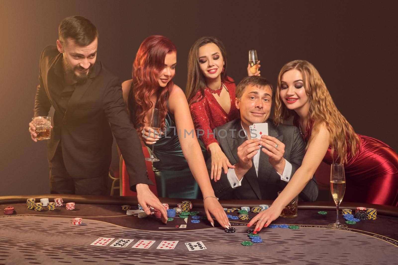 Funny colleagues are playing poker at casino. They are celebrating their win, smiling and looking vey excited while posing at the table against a dark smoke background in a ray of a spotlight. Cards, chips, money, alcohol, gambling, entertainment concept.