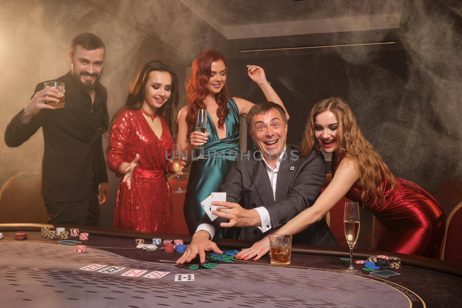 Enthusiastic buddies are playing poker at casino. They are celebrating their win, smiling and looking vey excited while posing at the table against a dark smoke background. Cards, chips, money, alcohol, gambling, entertainment concept.
