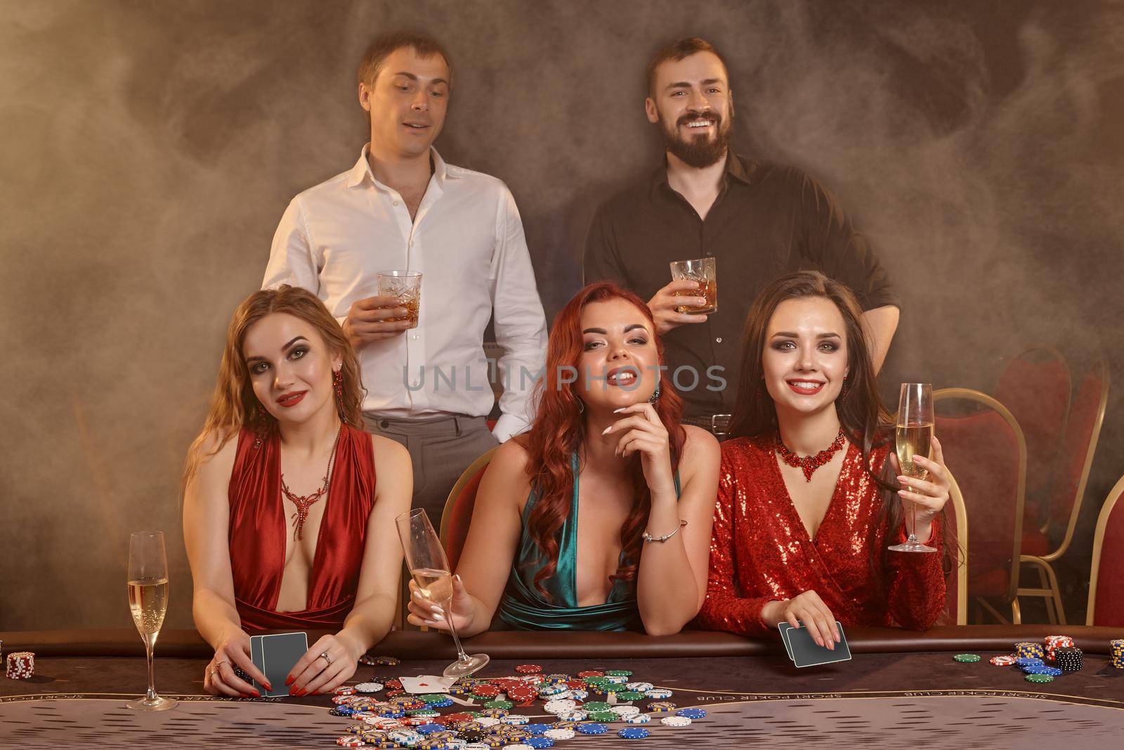 Enthusiastic buddies are playing poker at casino. They are celebrating their win, smiling and posing at the table against a dark smoke background. Cards, chips, money, alcohol, fortune, gambling, entertainment concept.