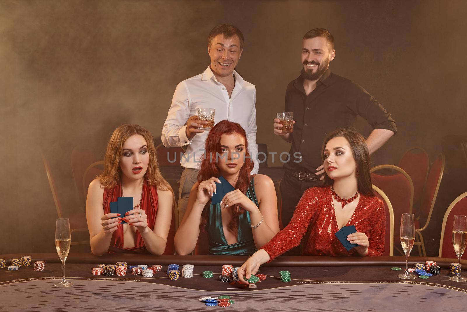 Concentrated friends are playing poker at casino. Golden youth are making bets waiting for a big win while posing at the table against a dark smoke background. Cards, chips, money, fortune, alcohol, gambling, entertainment concept.