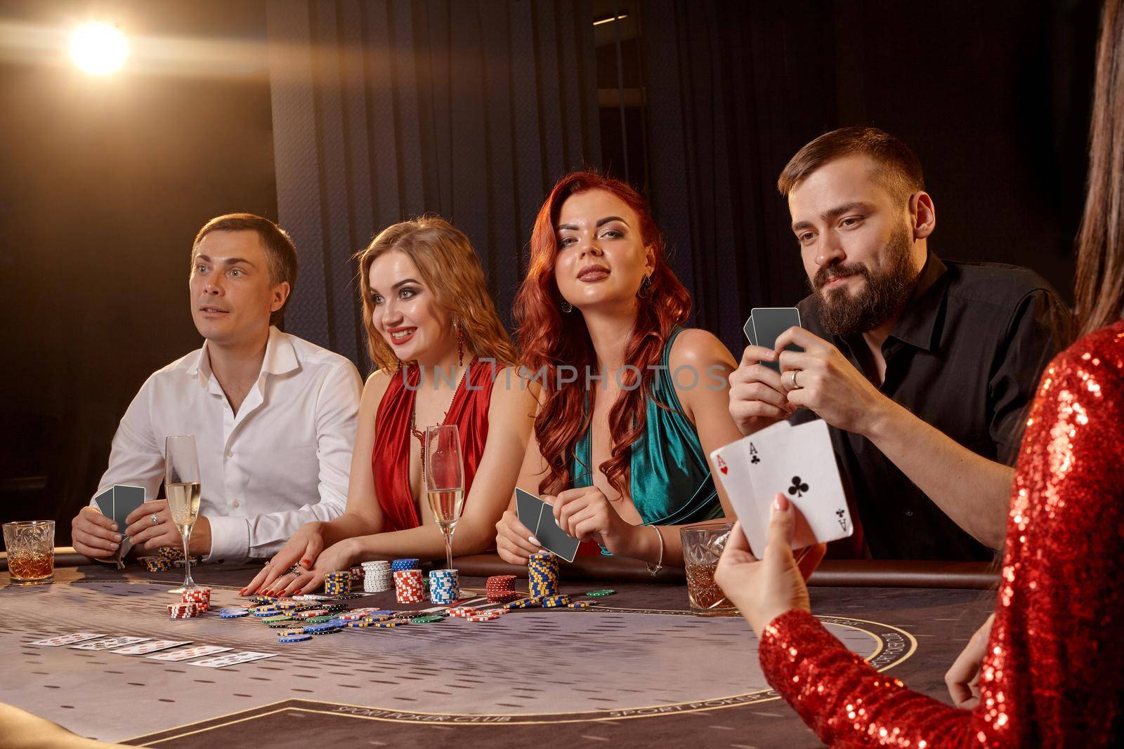 Enthusiastic companions are playing poker at casino. Youth are making bets waiting for a big win while posing at the table against a white spotlight on a dark smoke background. Cards, chips, money, alcohol, gambling, entertainment concept.