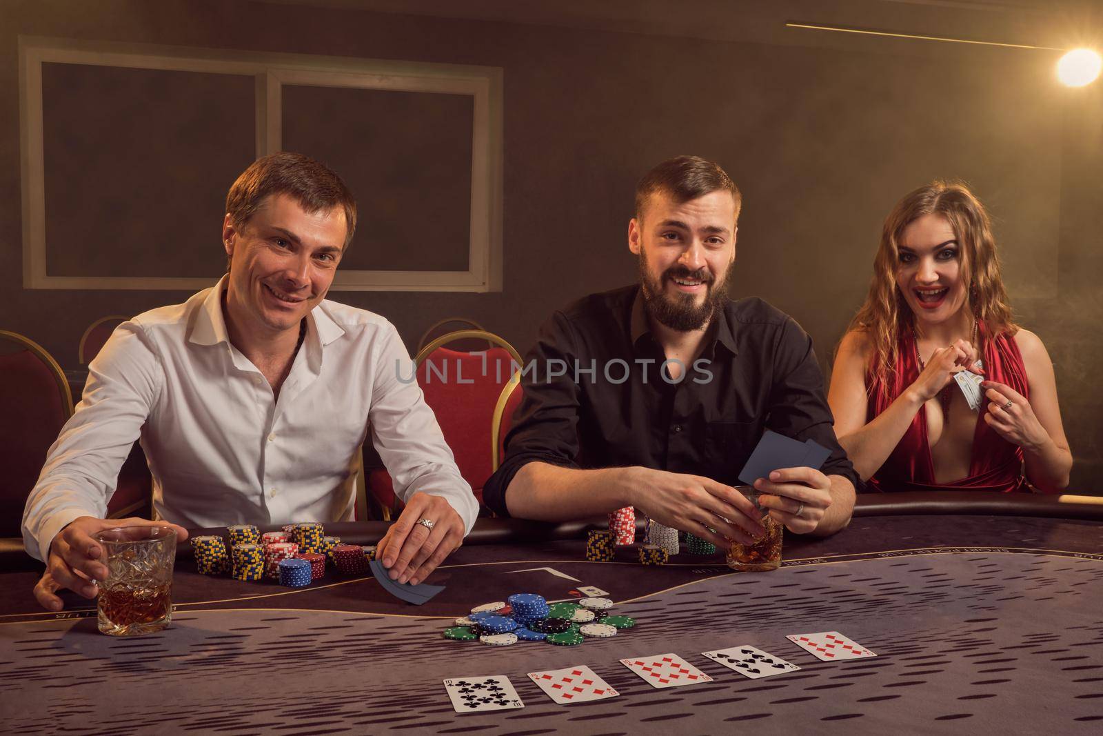 Two wealthy friends and beautiful female are playing poker at casino. They are making bets waiting for a big win, smiling and having a good time while posing sitting at the table against a yellow backlight on smoke background. Cards, chips, money, gambling, entertainment concept.