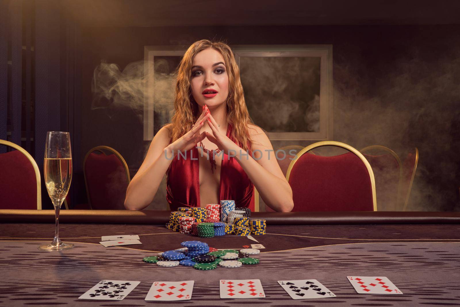 Attractive maiden with a long curly hair and perfect make-up, dressed in a sexy red dress. She is sitting at a gambling table, folded her hands and looking at the camera. Poker concept on a dark smoke background in a ray of a yellow spotlight. Casino.