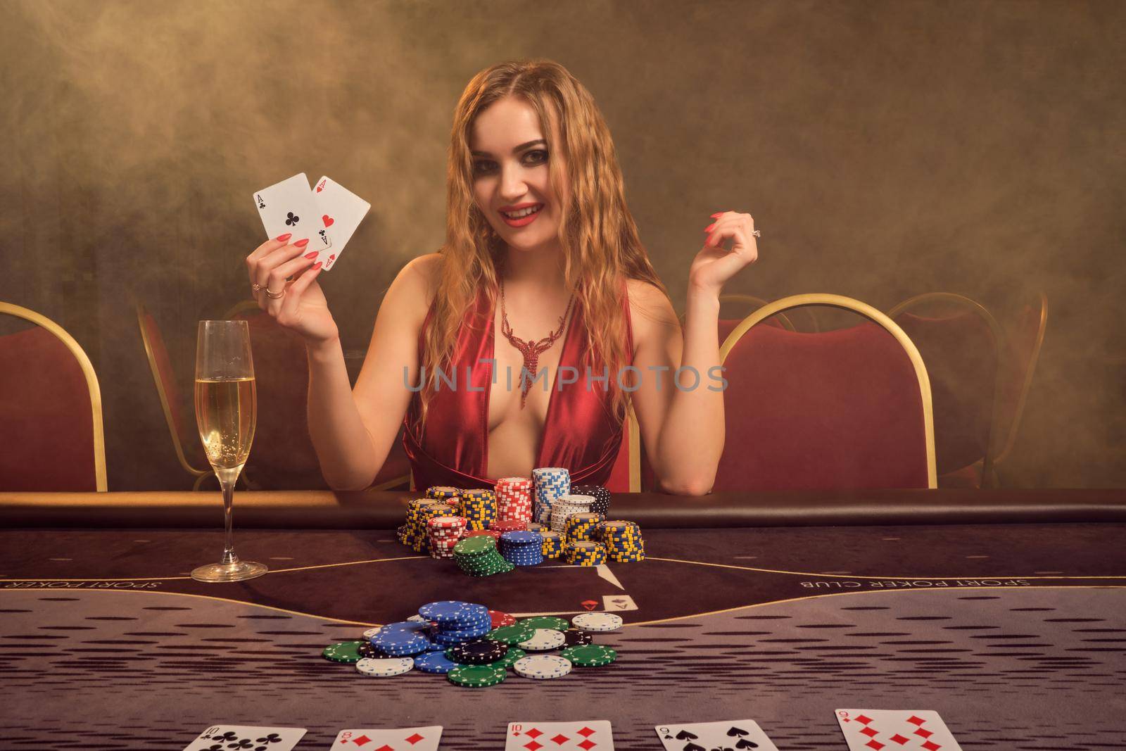 Alluring lady with a long curly hair and perfect make-up, dressed in a sexy red dress. She is sitting at a gambling table with two playing cards in her hands and smiling. Poker concept on a dark smoke background in a ray of a yellow spotlight. Casino.