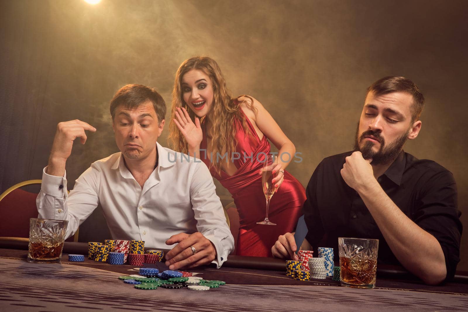 Two old friends and attractive woman are playing poker at casino. They are making bets waiting for a big win while posing at the table against a yellow backlight on smoke background. Cards, chips, money, gambling, entertainment concept.