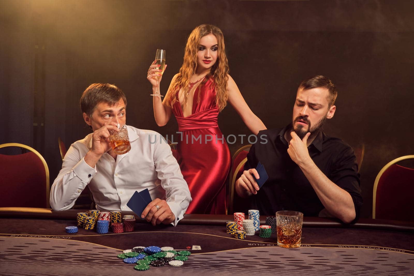 Two good-looking friends and sexy woman are playing poker at casino. They are making bets waiting for a big win while posing at the table against a yellow backlight on smoke background. Cards, chips, money, gambling, entertainment concept.