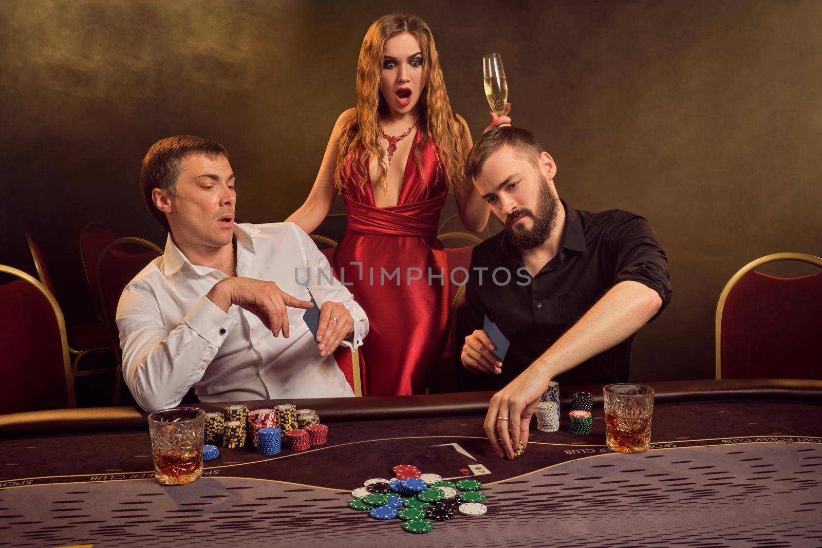 Two old friends and attractive maiden are playing poker at casino. They are making bets waiting for a big win and looking wondered while posing at the table against a yellow backlight on smoke background. Cards, chips, money, gambling, entertainment concept.