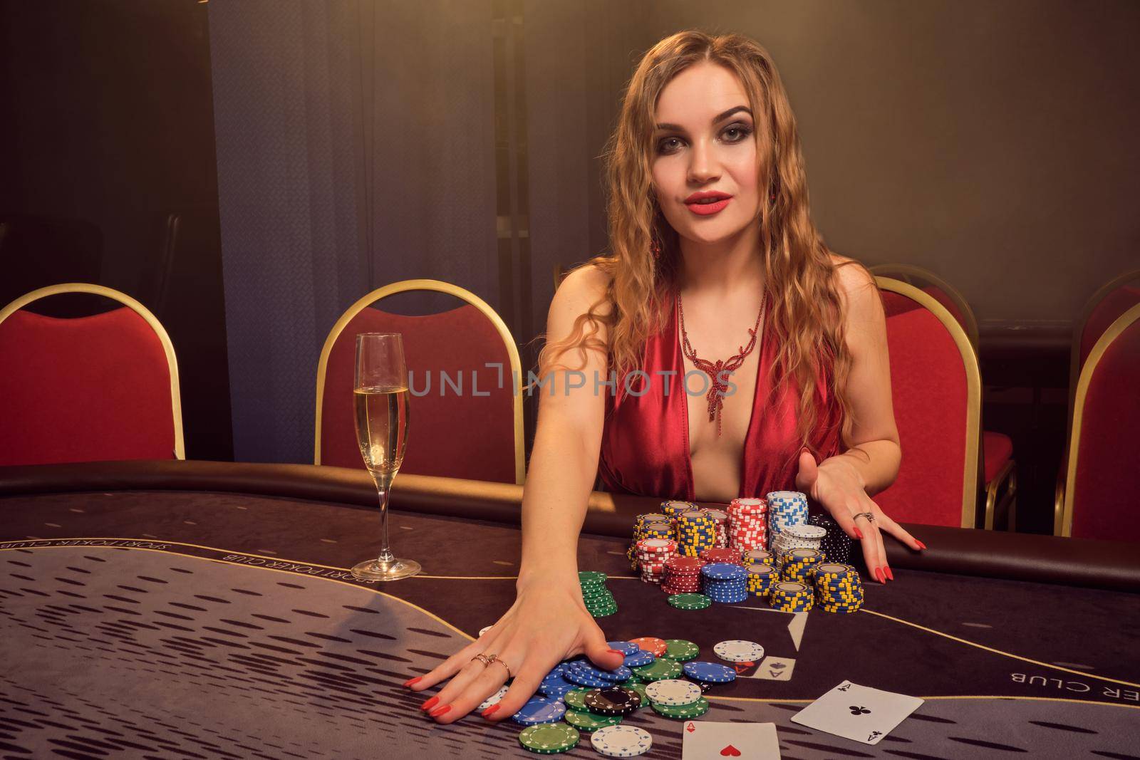 Beautiful female with a long curly hair and perfect make-up, dressed in a sexy red dress. She is rejoicing her win while sitting at a gambling table and looking at the camera. Poker concept on a dark smoke background in a ray of a yellow spotlight. Casino.