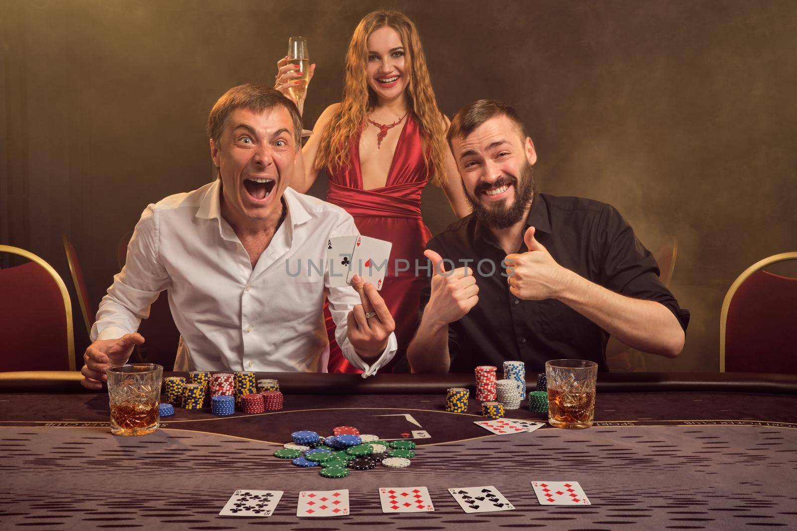 Two wealthy friends and beautiful maiden are playing poker at casino. They are rejoicing their win, smiling and looking at the camera while posing at the table against a yellow backlight on smoke background. Cards, chips, money, gambling, entertainment concept.