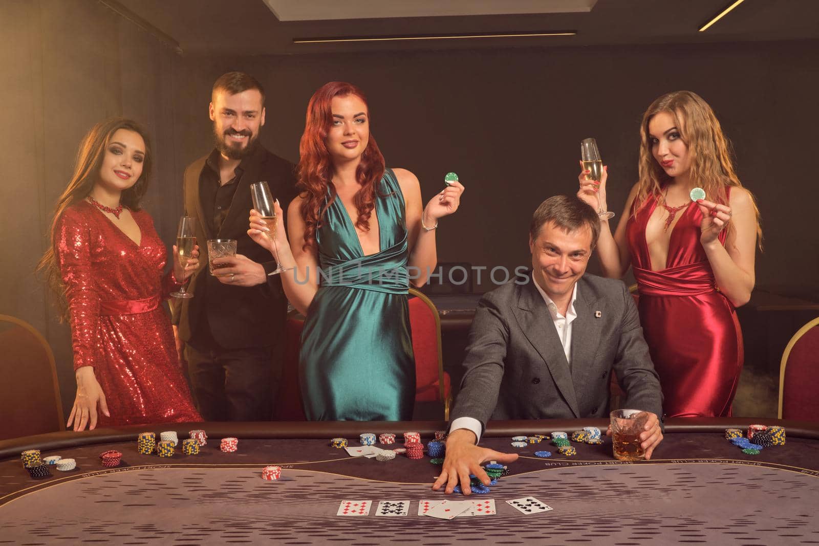 Happy partners are playing poker at casino. They are celebrating their win, smiling and looking vey excited while posing at the table against a dark smoke background in a ray of a spotlight. Cards, chips, money, alcohol, gambling, entertainment concept.