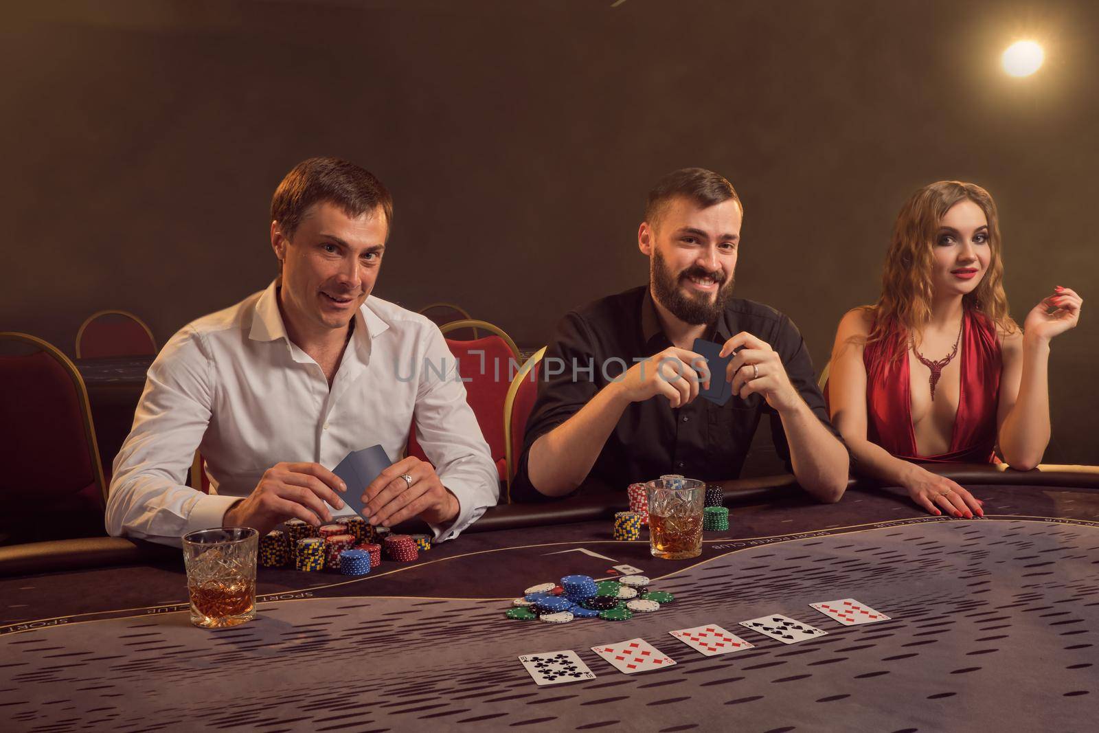 Two wealthy friends and beautiful woman are playing poker at casino. They are making bets waiting for a big win, smiling and looking at the camera while posing sitting at the table against a yellow backlight on dark background. Cards, chips, money, gambling, entertainment concept.