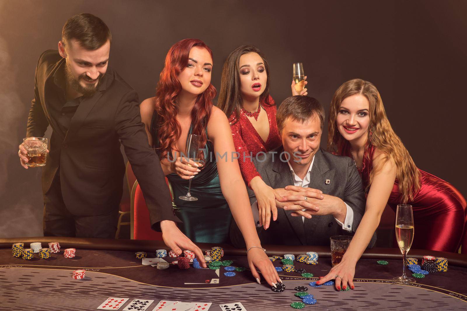 Funny partners are playing poker at casino. They are celebrating their win, smiling and looking vey excited while posing at the table against a dark smoke background. Cards, chips, money, alcohol, gambling, entertainment concept.