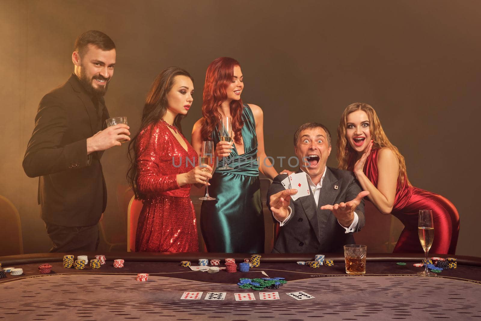 Enthusiastic partners are playing poker at casino. They are celebrating their win, smiling and looking vey excited while posing at the table against a dark smoke background in a ray of a spotlight. Cards, chips, money, alcohol, gambling, entertainment concept.
