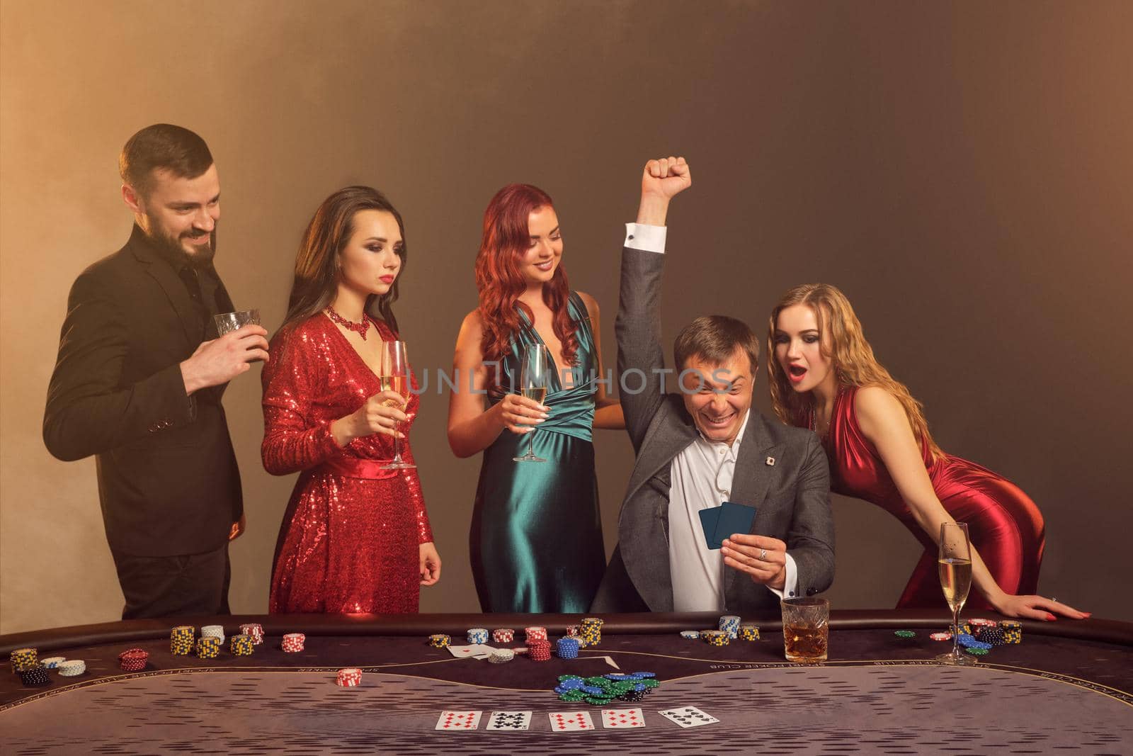 Funny buddies are playing poker at casino. They are celebrating their win, smiling and looking vey excited while posing at the table against a dark smoke background in a ray of a spotlight. Cards, chips, money, alcohol, gambling, entertainment concept.