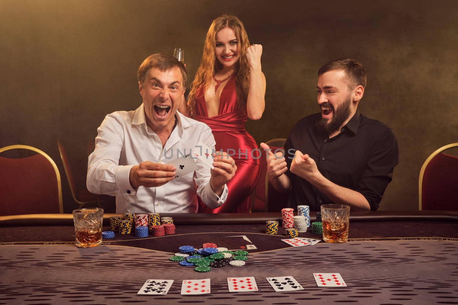 Two athletic friends and charming lady are playing poker at casino. They are rejoicing their win and showing two aces while posing at the table against a yellow backlight on smoke background. Cards, chips, money, gambling, entertainment concept.