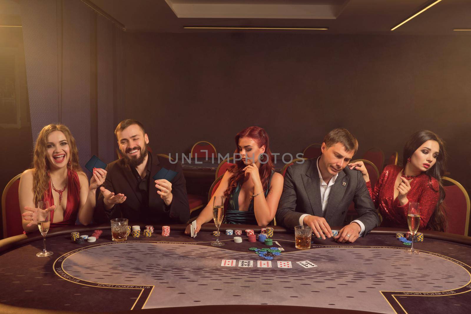 Merry colleagues are playing poker at casino. They are making bets waiting for a big win while posing at the table against a dark smoke background in a ray of a spotlight. Cards, chips, money, alcohol, gambling, entertainment concept.
