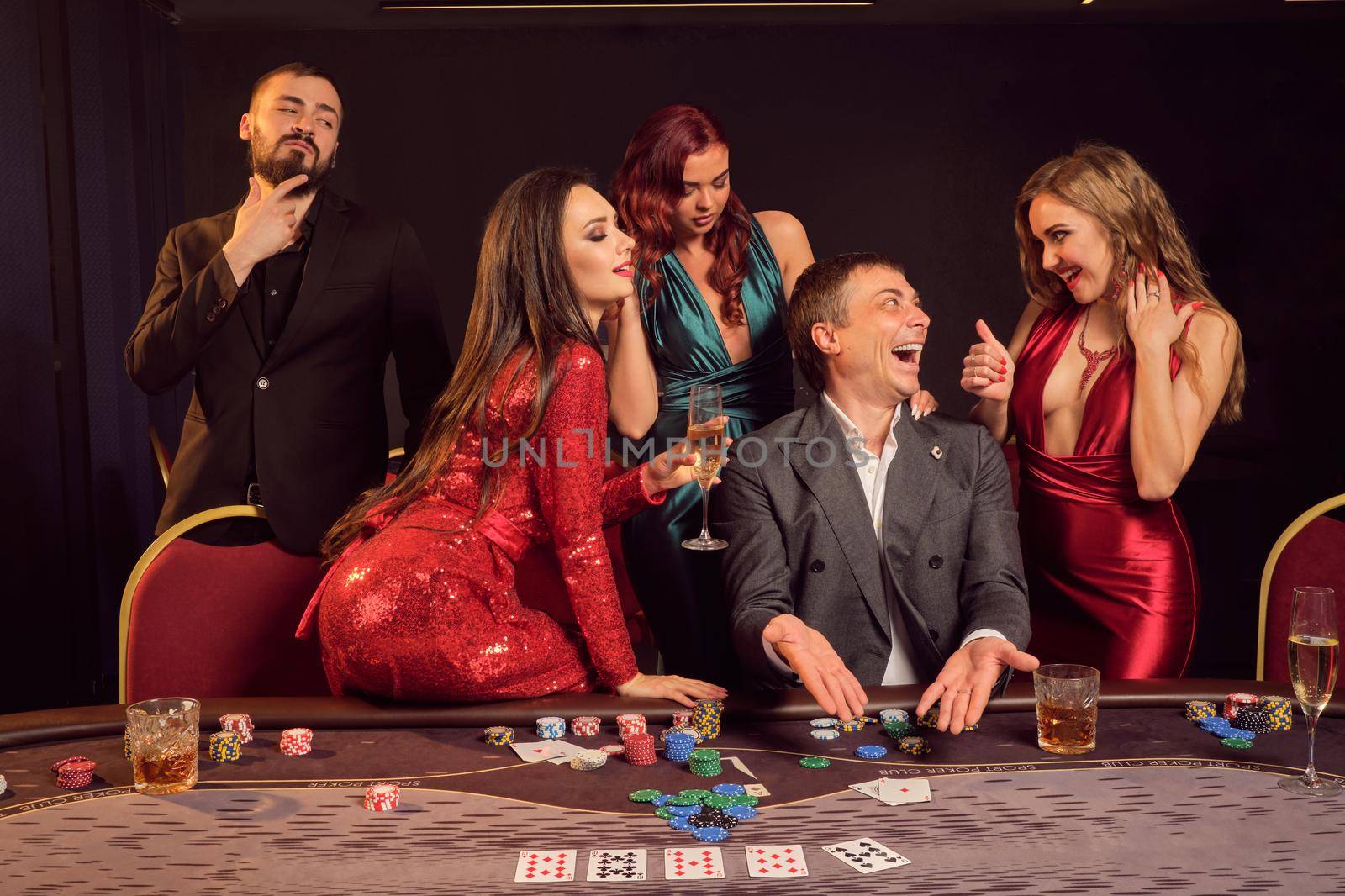 Optimistic friends are playing poker at casino. They are celebrating their win, smiling and looking vey excited while posing at the table against a dark background. Cards, chips, money, alcohol, gambling, entertainment concept.